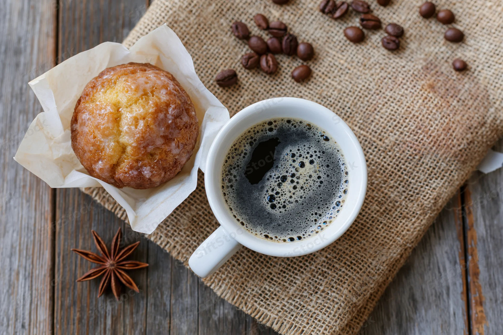White ceramic cup of black coffee with foam beside a glazed donut on burlap over wooden table