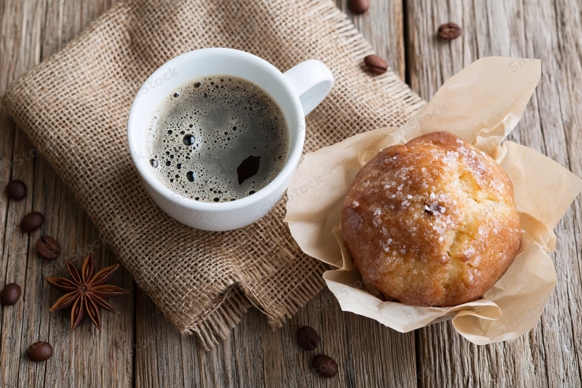 White ceramic cup of black coffee with foam beside a sugar-dusted pastry on a wooden table with burlap cloth and coffee beans