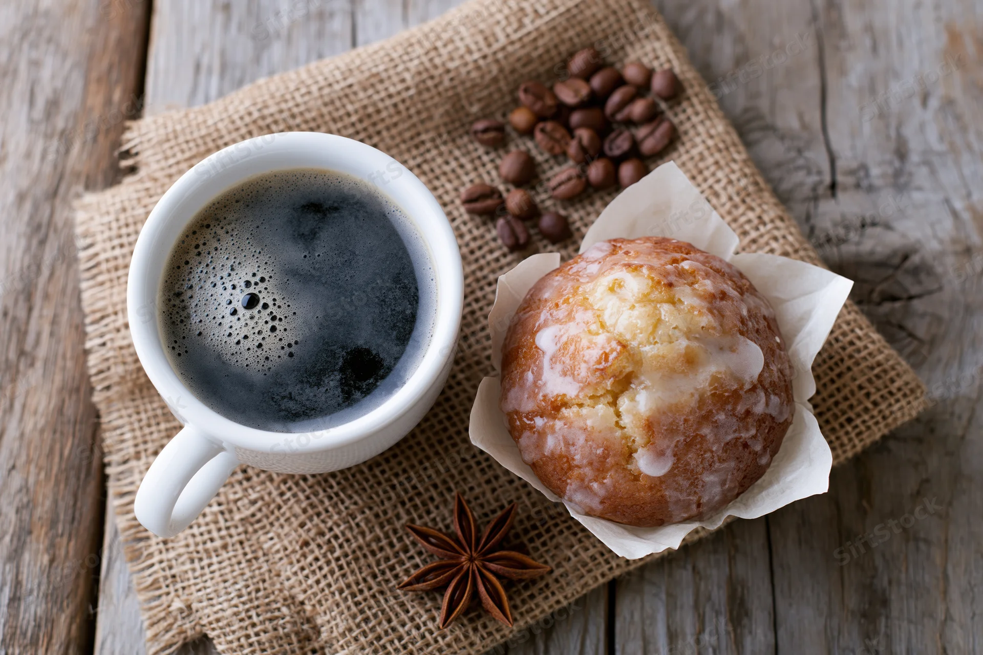 White ceramic cup of black coffee with foam beside a glazed muffin on burlap over wooden table