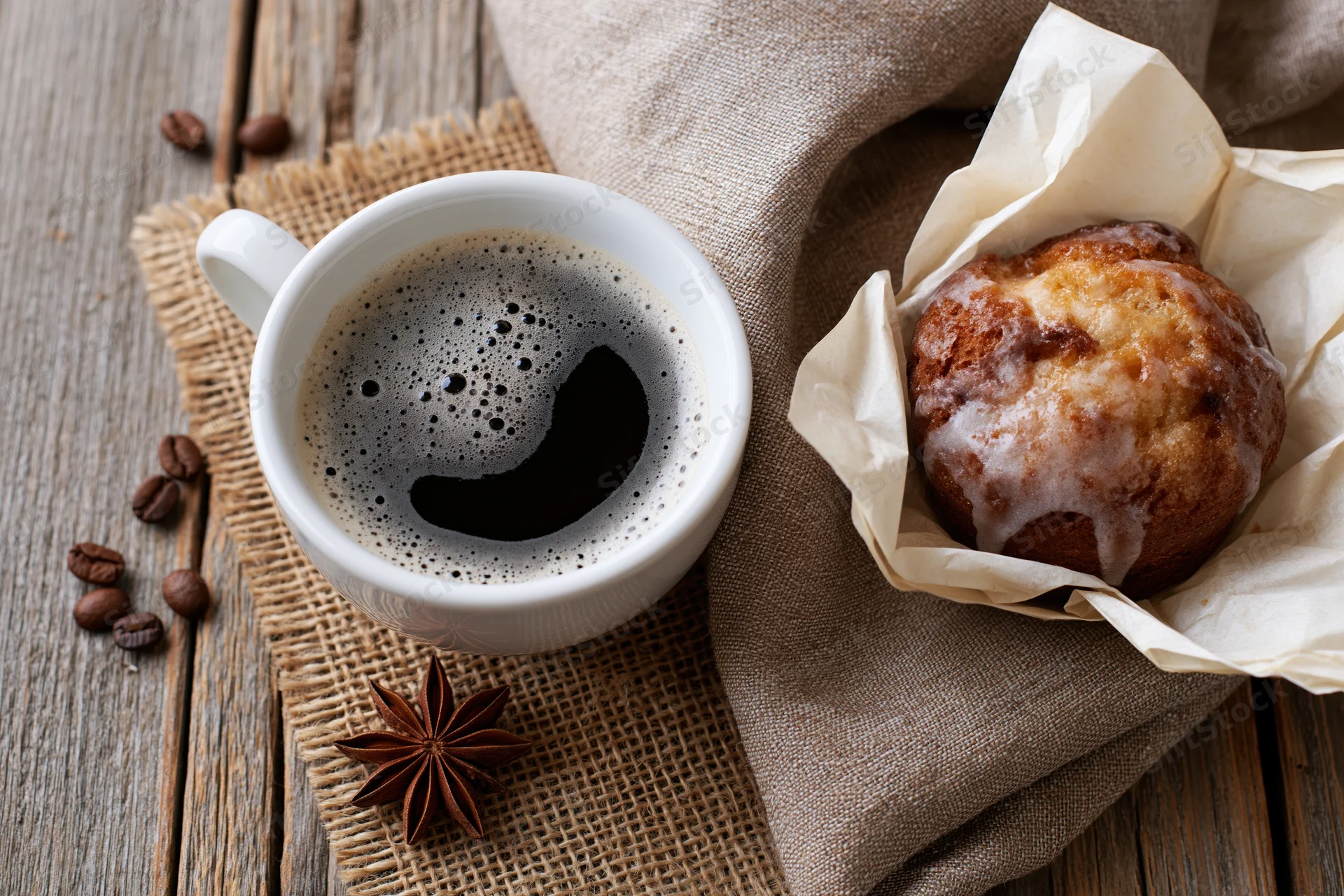 White ceramic cup of black coffee with foam beside a glazed muffin on burlap and wooden table