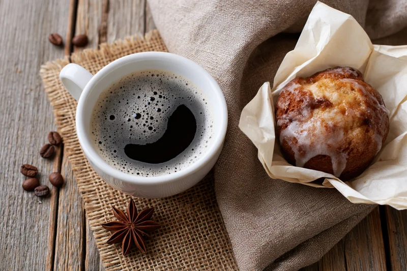 White ceramic cup of black coffee with foam beside a glazed muffin on burlap and wooden table