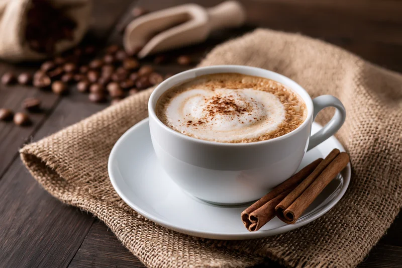White ceramic cup of cappuccino with heart-shaped foam and cinnamon sticks on saucer over burlap on wooden table
