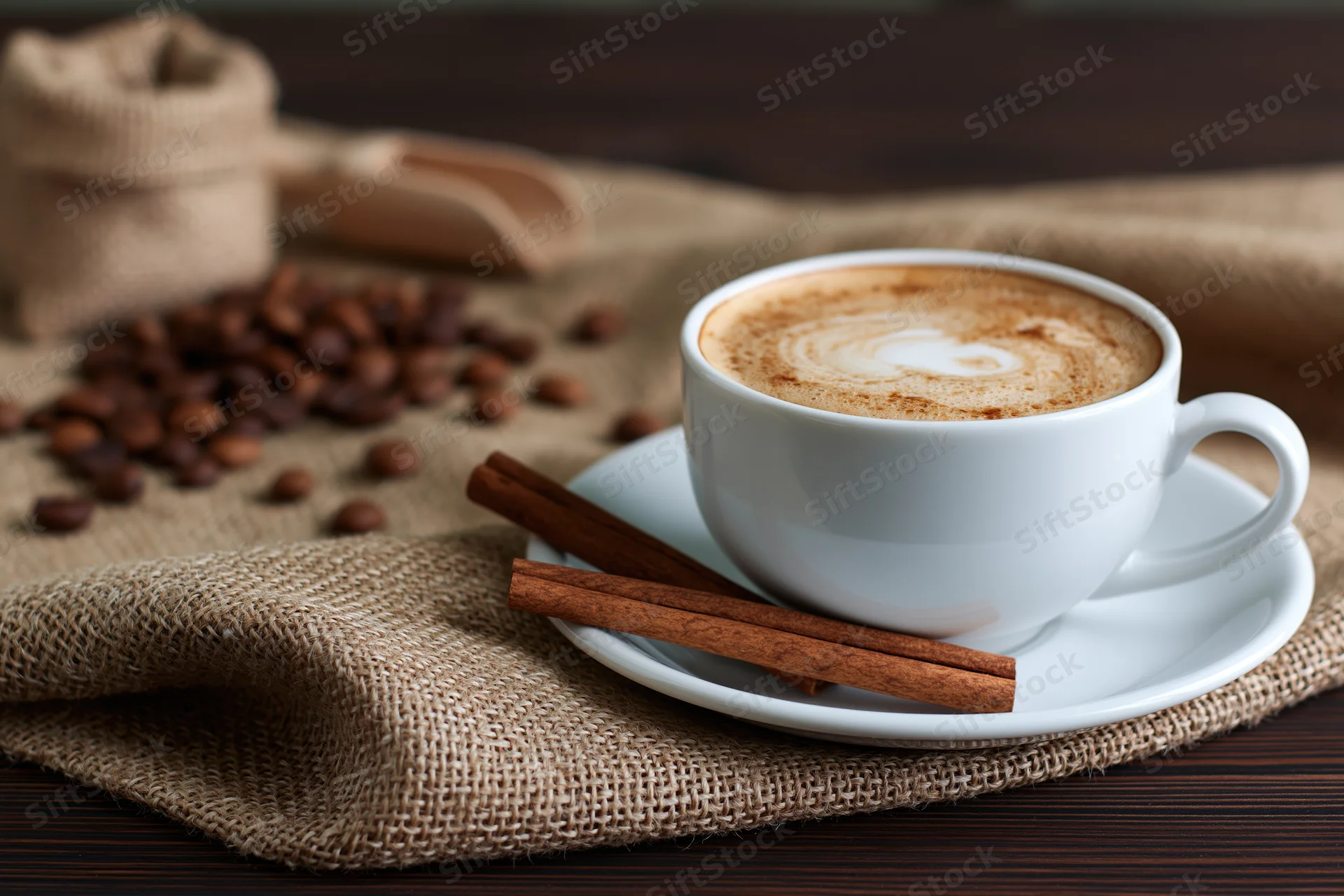 White ceramic cup of cappuccino with latte art and cinnamon sticks on a burlap cloth over dark wood table