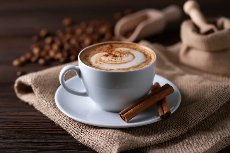 White ceramic cup of cappuccino with latte art and cinnamon sticks on a saucer over burlap cloth on wooden table