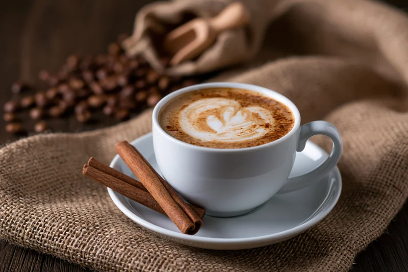 White ceramic cup of cappuccino with latte art heart on saucer beside cinnamon sticks and coffee beans on burlap cloth