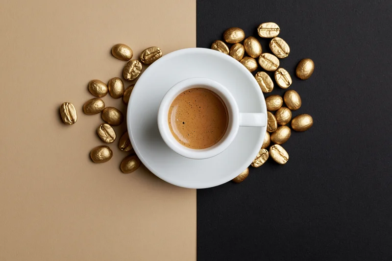 White ceramic espresso cup filled with coffee on saucer surrounded by golden coffee beans on split beige and black background