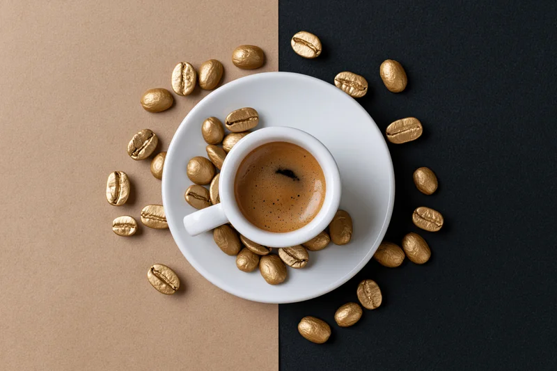 White ceramic espresso cup filled with coffee on saucer surrounded by golden coffee beans on split beige and black background