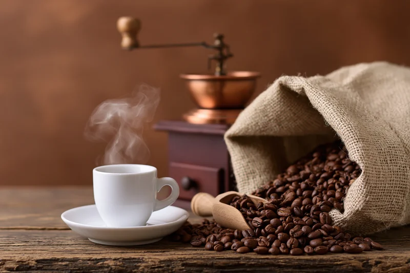 Steaming espresso cup beside spilled coffee beans and manual grinder on rustic wooden table