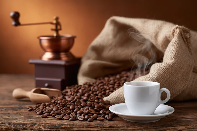 Steaming white ceramic espresso cup on wooden table beside coffee beans and manual grinder with burlap sack