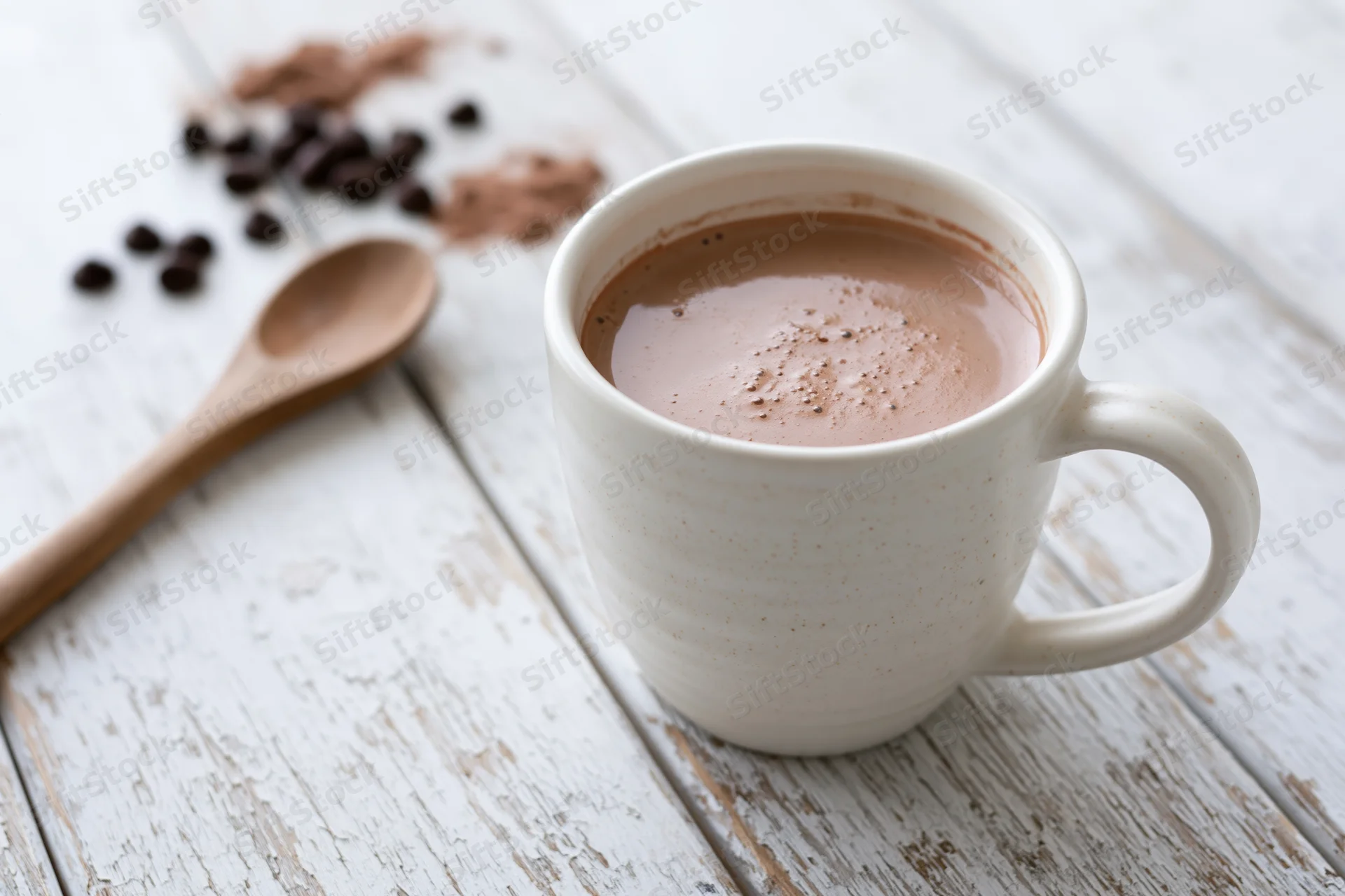 White ceramic mug filled with hot chocolate on a rustic wooden table with scattered coffee beans and a wooden spoon nearby