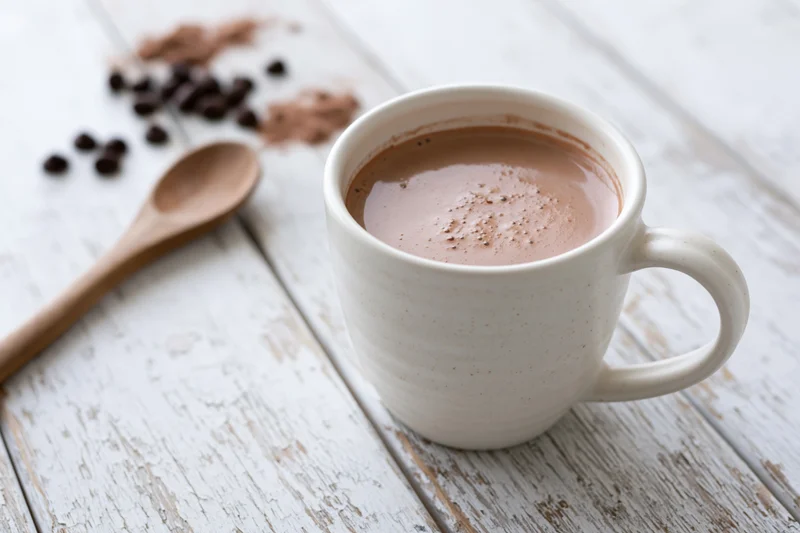 White ceramic mug filled with hot chocolate on a rustic wooden table with scattered coffee beans and a wooden spoon nearby