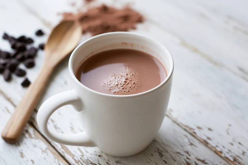 White ceramic mug filled with hot chocolate on a rustic wooden table with chocolate chips and cocoa powder nearby