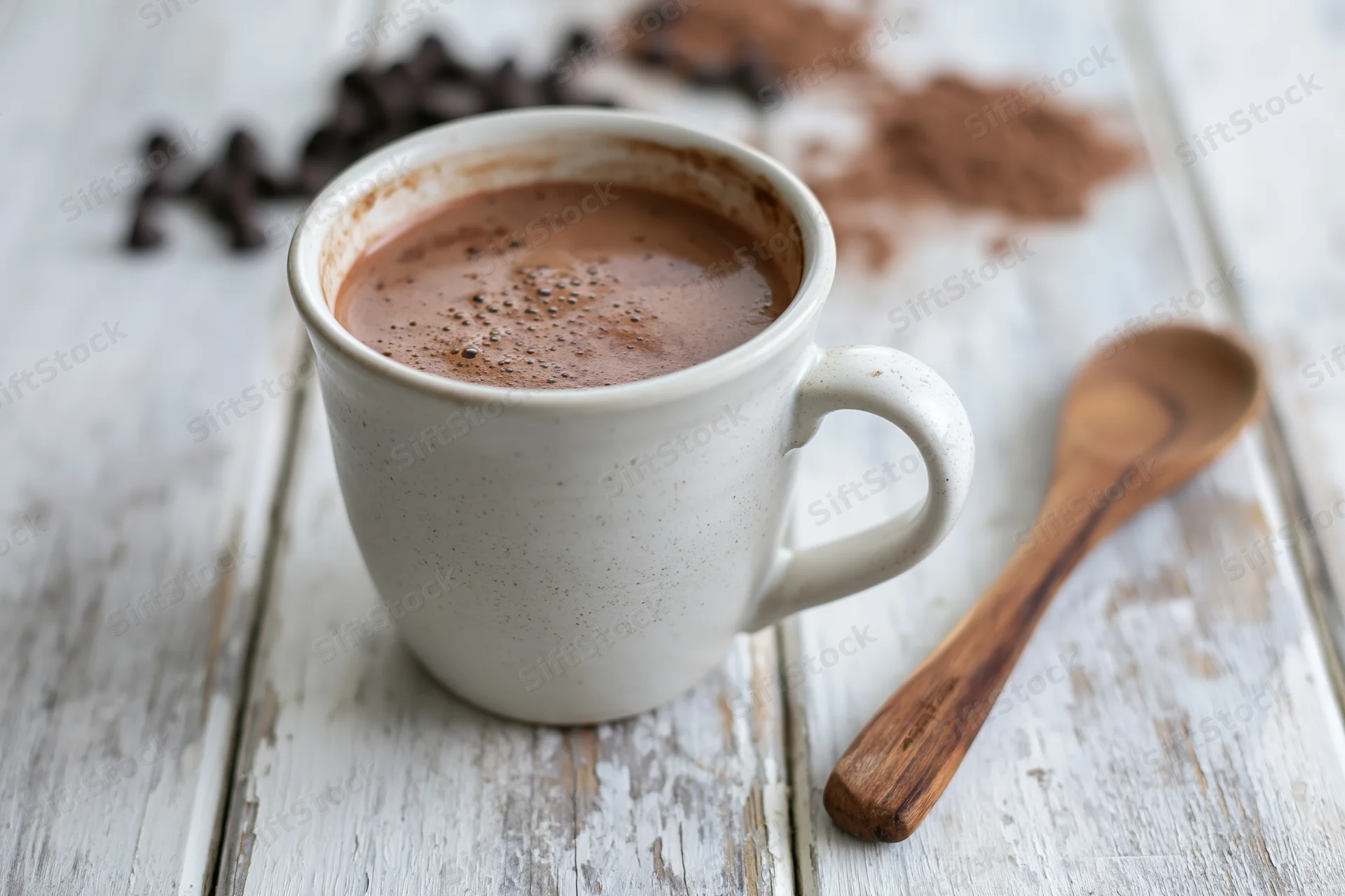 White ceramic mug filled with hot chocolate on a rustic wooden table with a wooden spoon and cocoa powder nearby