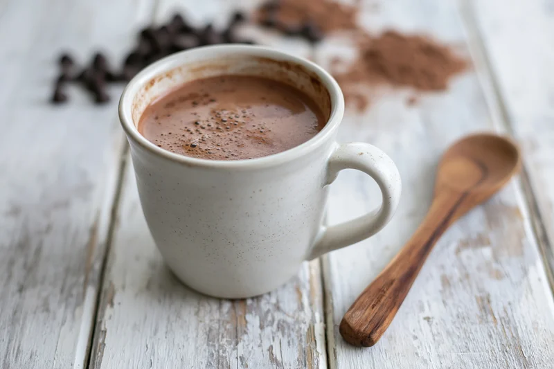 White ceramic mug filled with hot chocolate on a rustic wooden table with a wooden spoon and cocoa powder nearby
