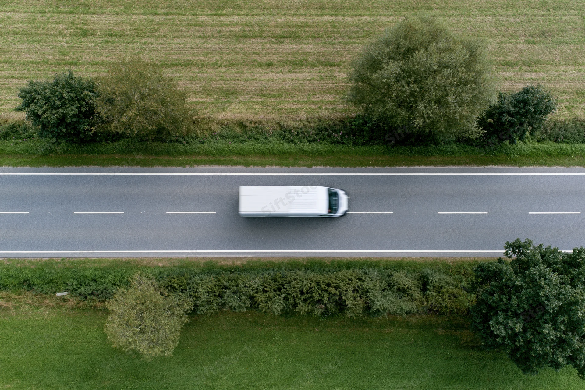 White delivery van driving on a straight two-lane rural road surrounded by green fields and trees, viewed from above