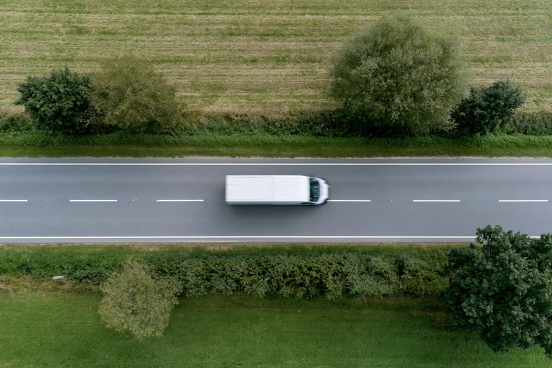 White delivery van driving on a straight two-lane rural road surrounded by green fields and trees, viewed from above