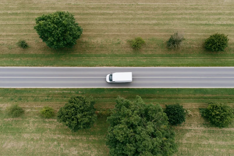 White delivery van driving on straight rural road surrounded by fields and trees from above