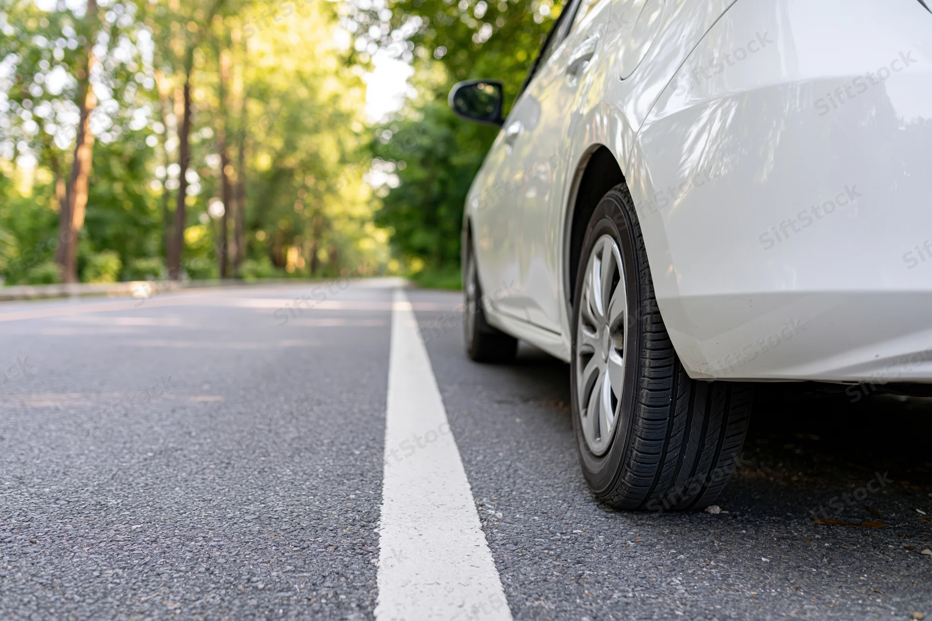 White hatchback parked on asphalt road beside white lane line with trees in background