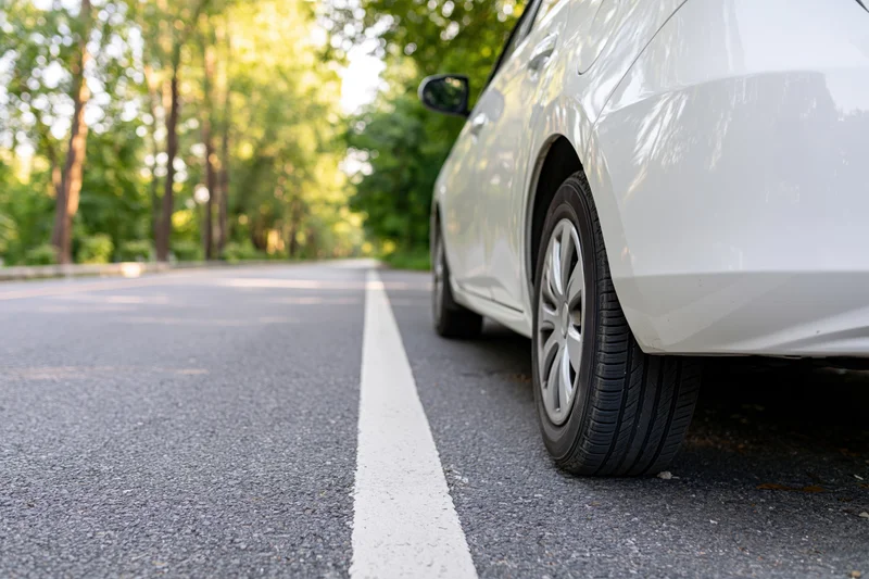 White hatchback parked on asphalt road beside white lane line with trees in background