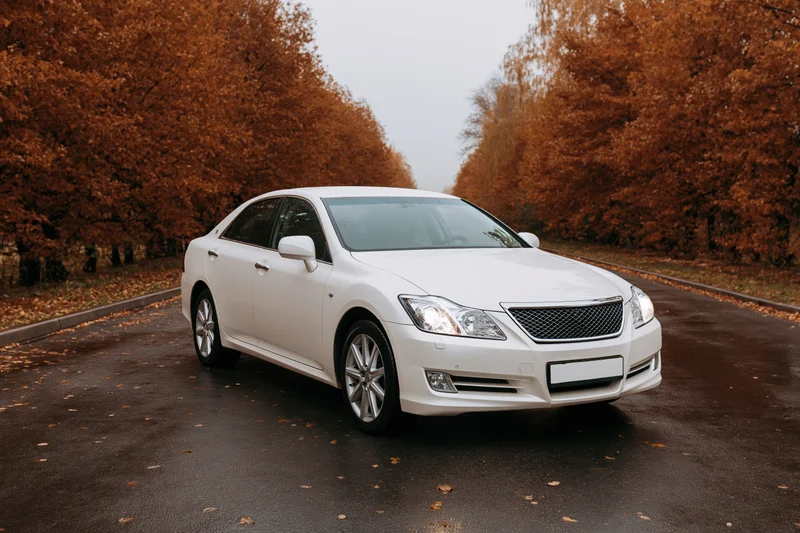 White luxury sedan parked on wet asphalt road with headlights on surrounded by autumn trees