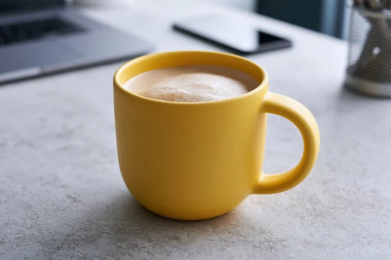 Yellow ceramic mug filled with frothy cappuccino on a gray desk beside a laptop and smartphone