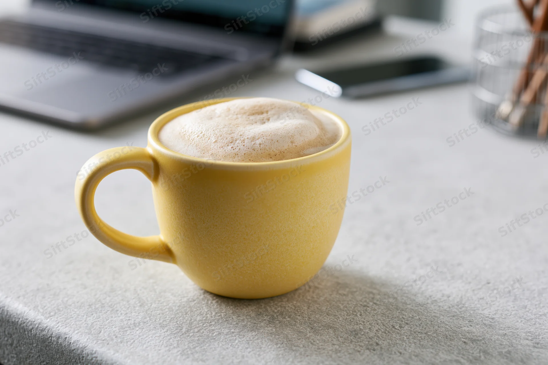 Yellow ceramic mug filled with frothy cappuccino on a gray desk beside a laptop and smartphone