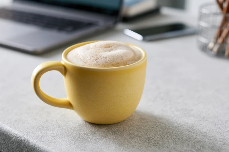 Yellow ceramic mug filled with frothy cappuccino on a gray desk beside a laptop and smartphone