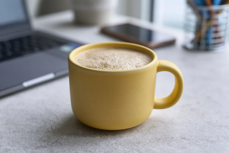Yellow ceramic mug filled with frothy cappuccino on a desk beside a laptop and smartphone