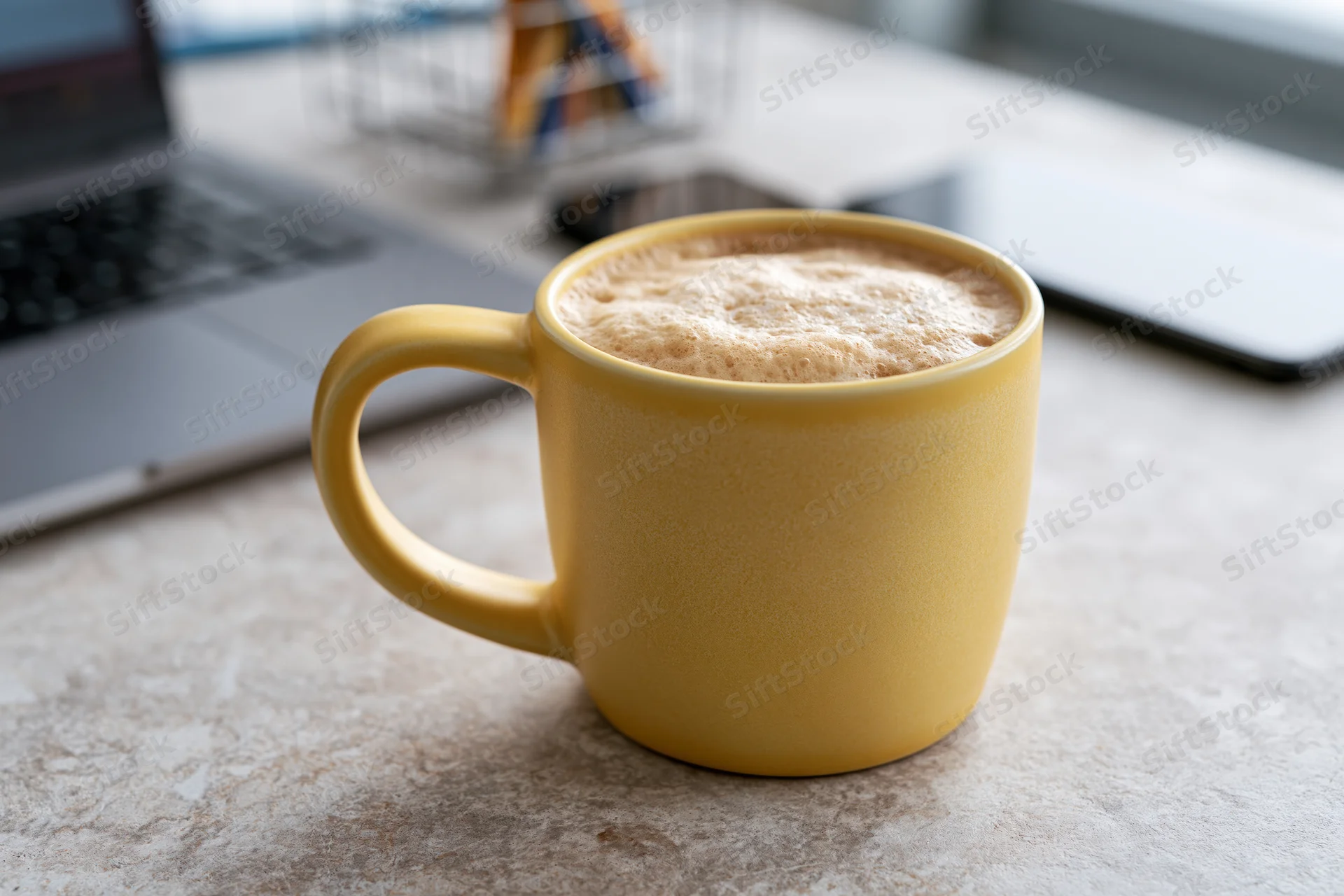 Yellow ceramic mug filled with frothy cappuccino on a desk beside a laptop