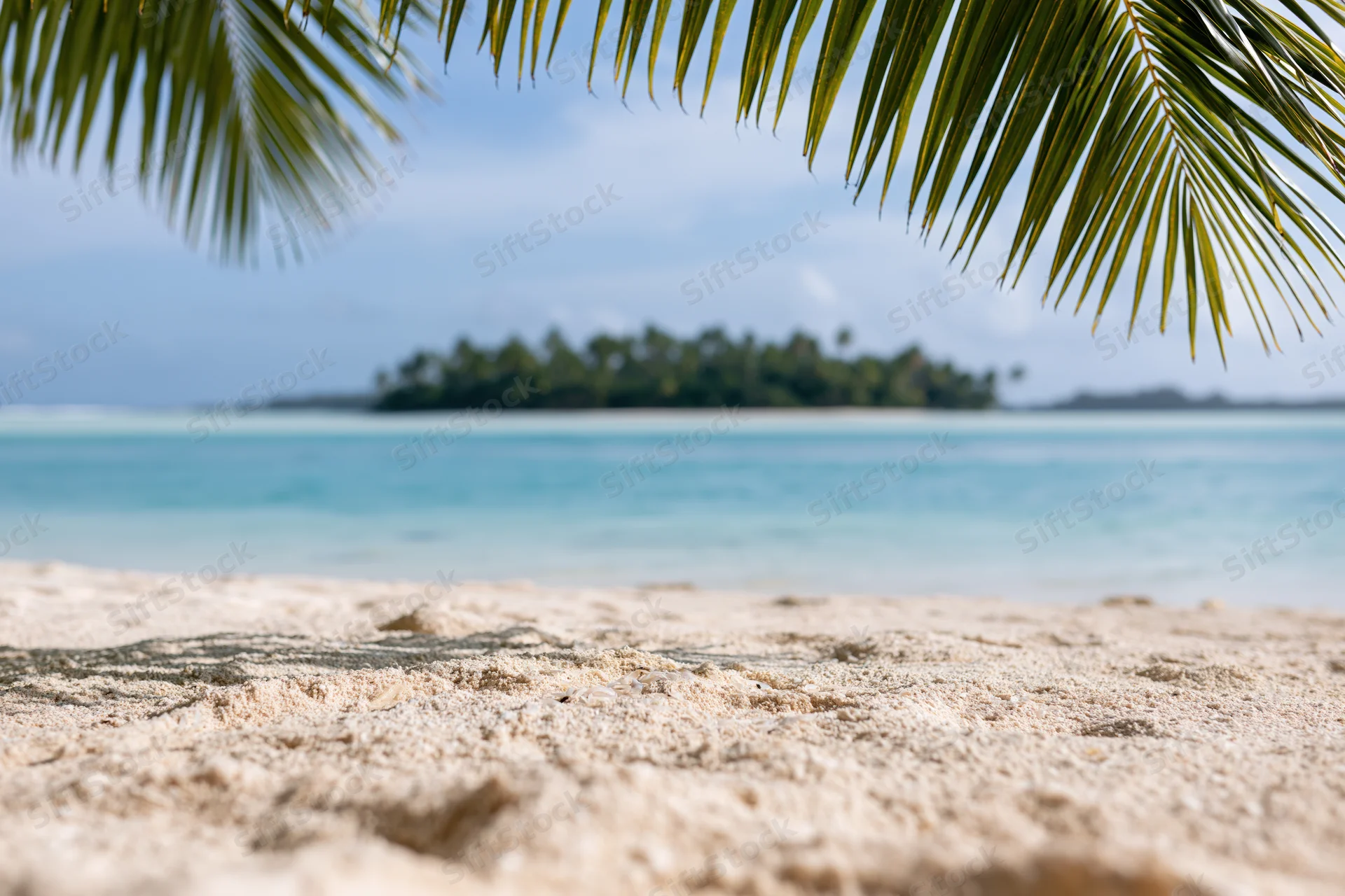 Close-up of sandy beach under palm fronds with distant tropical island across turquoise water