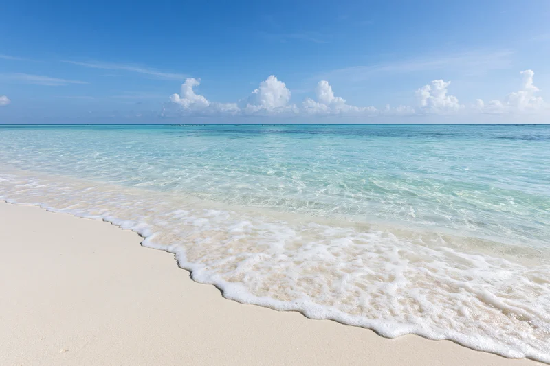 Gentle waves rolling onto white sand beach under clear blue sky with scattered clouds