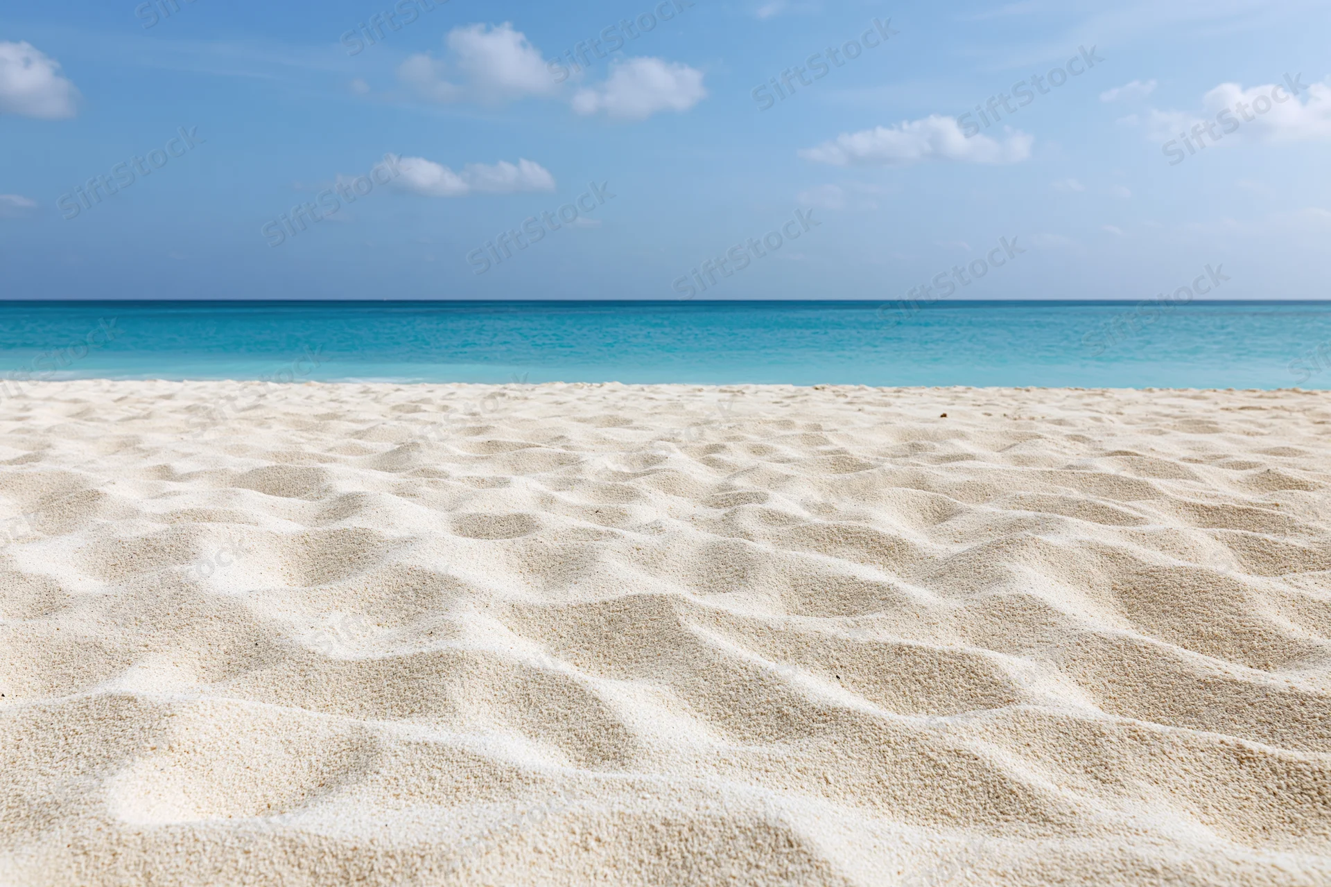 Textured white sand along a calm turquoise ocean under a blue sky with scattered clouds