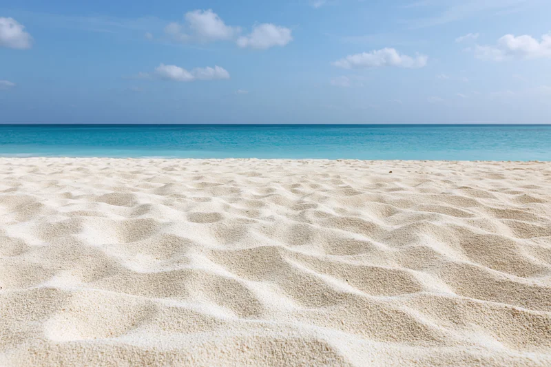 Textured white sand along a calm turquoise ocean under a blue sky with scattered clouds