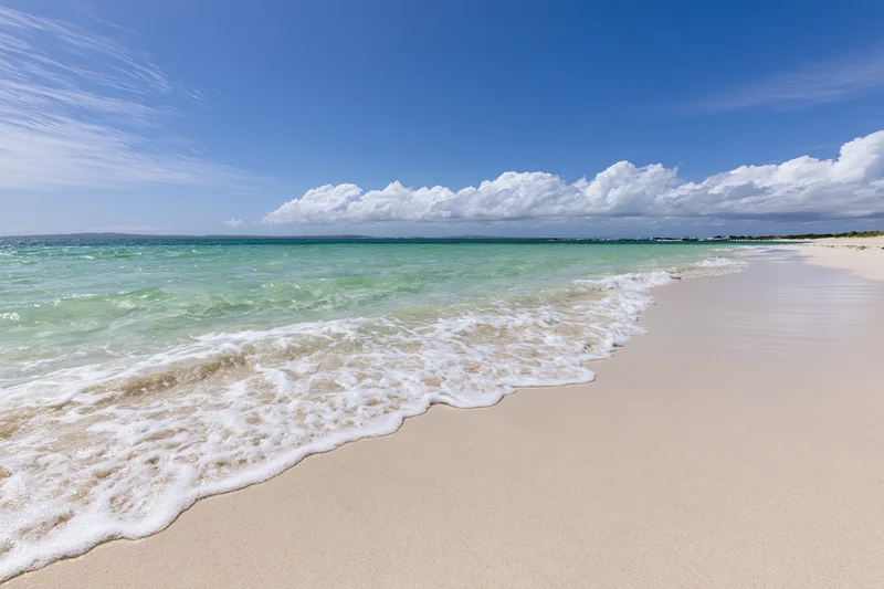 Gentle waves rolling onto a sandy beach under a clear blue sky with scattered clouds