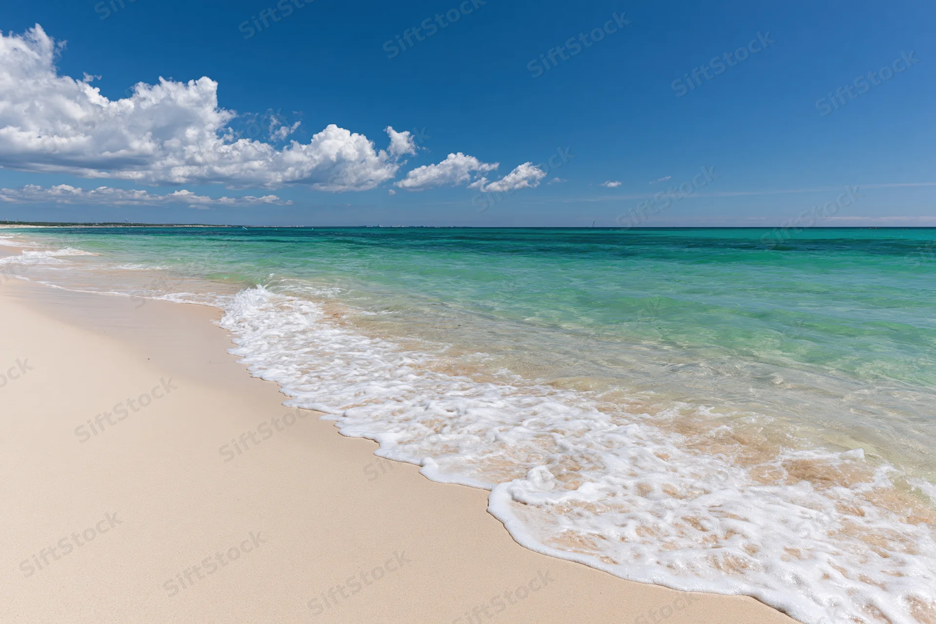 Gentle waves rolling onto a sandy beach under a clear blue sky with scattered clouds