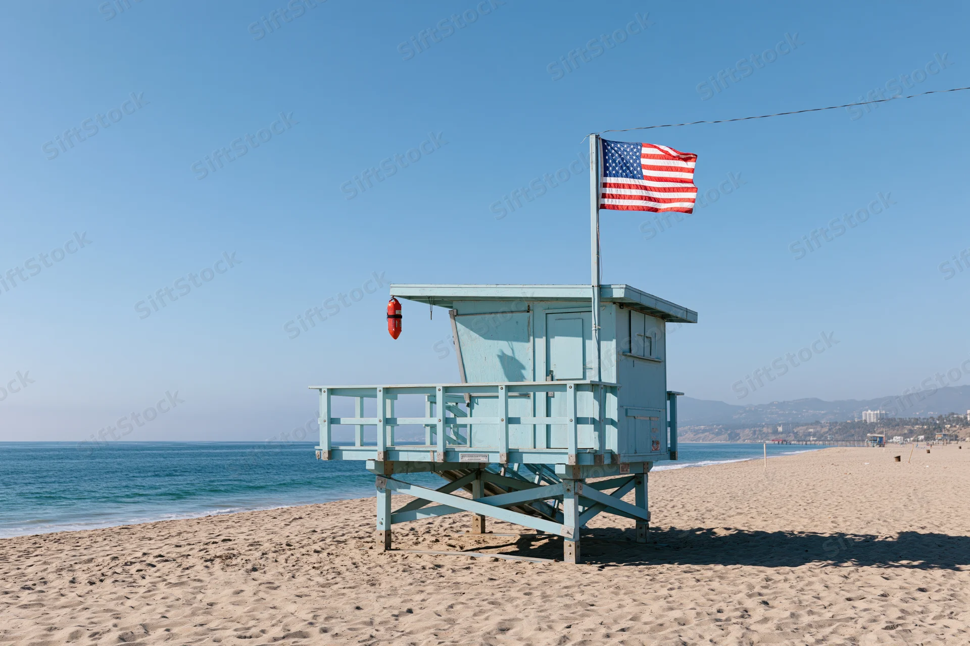 Light blue lifeguard tower on sandy beach with american flag flying, overlooking ocean under clear blue sky