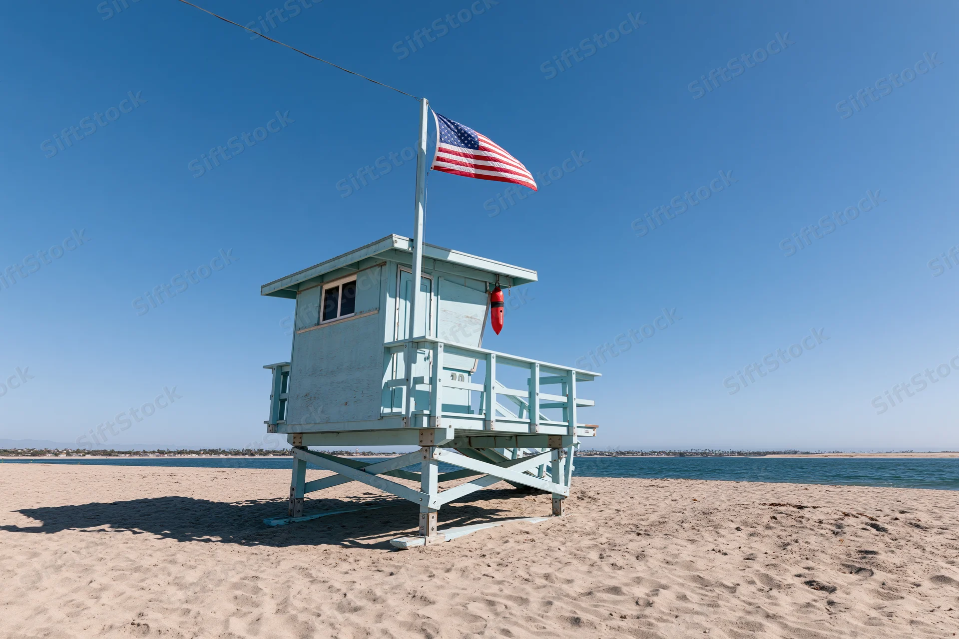 Light blue wooden lifeguard tower on sandy beach with american flag flying under clear blue sky