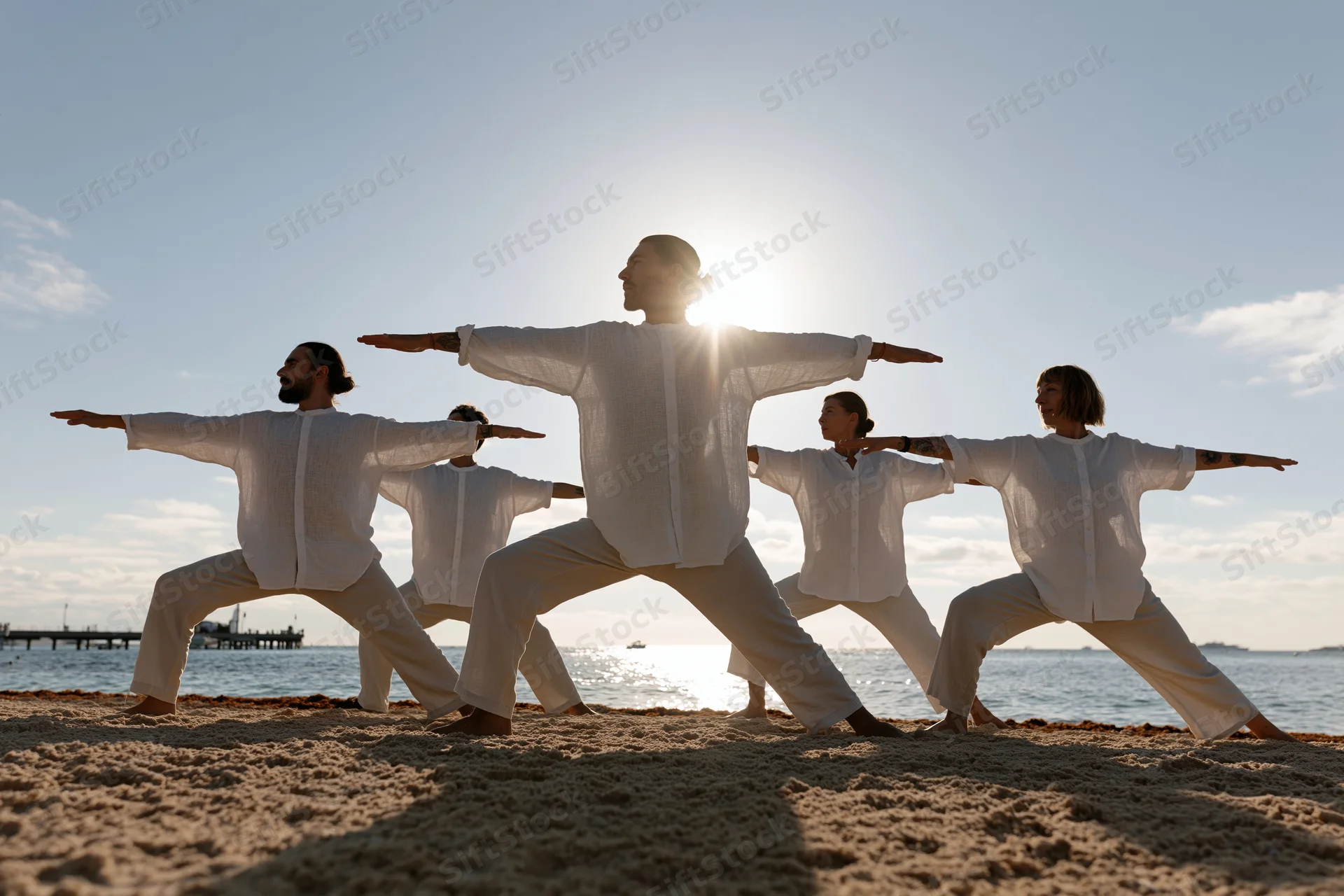 Group practicing yoga in warrior pose on sandy beach at sunrise with ocean in background