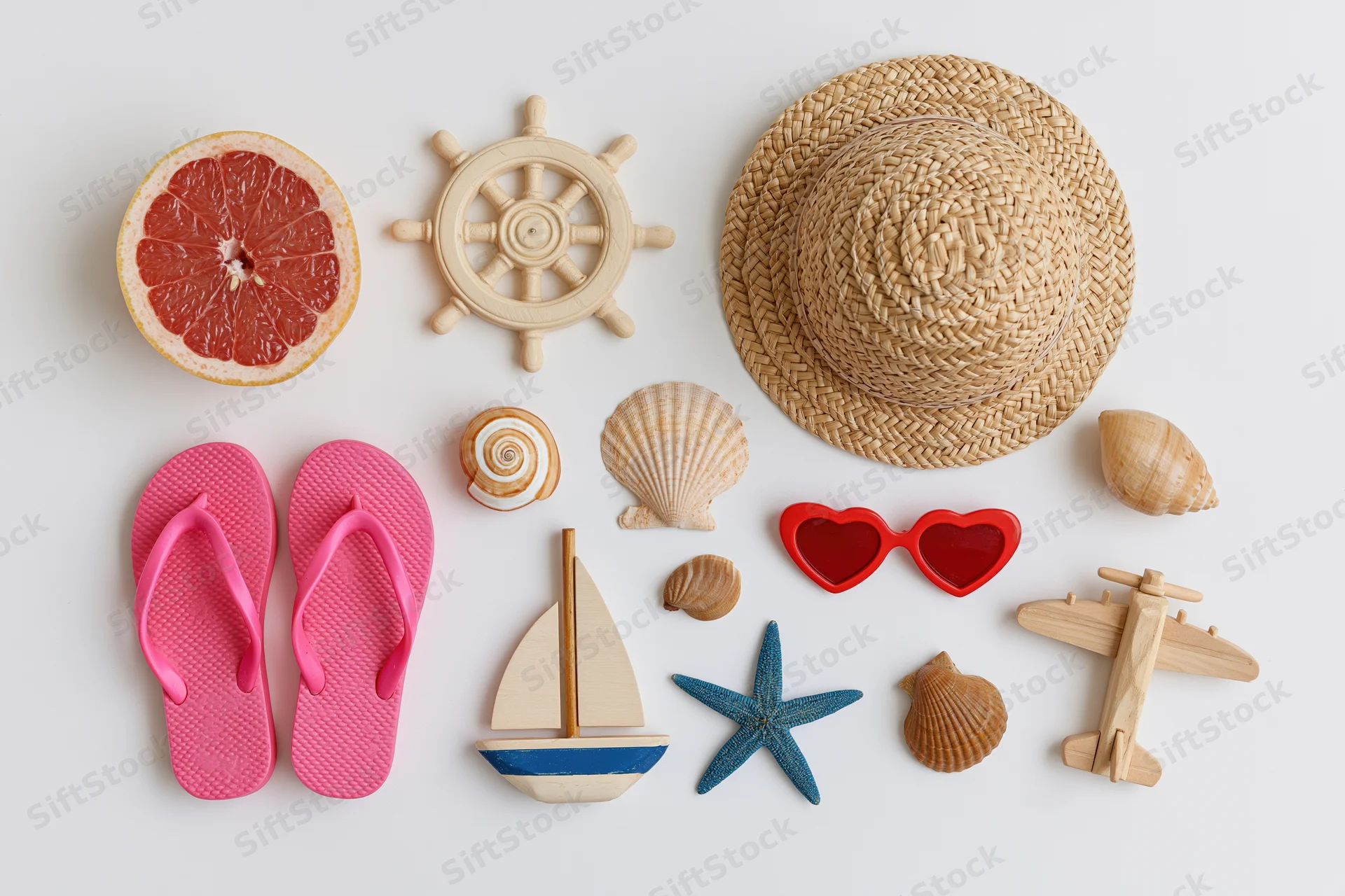 Summer vacation items arranged on white surface including flip flops, straw hat, sunglasses, seashells, grapefruit, wooden toys, and nautical decor