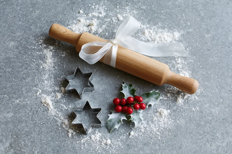 Wooden rolling pin tied with white ribbon, star-shaped cookie cutters and holly with red berries on a gray floured surface