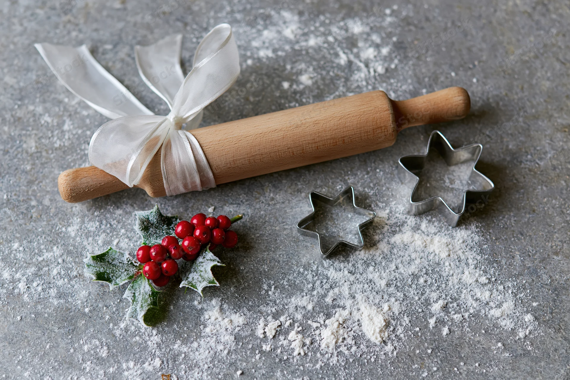Wooden rolling pin tied with white ribbon and star-shaped cookie cutters on a floured gray countertop with holly berries, for holiday baking