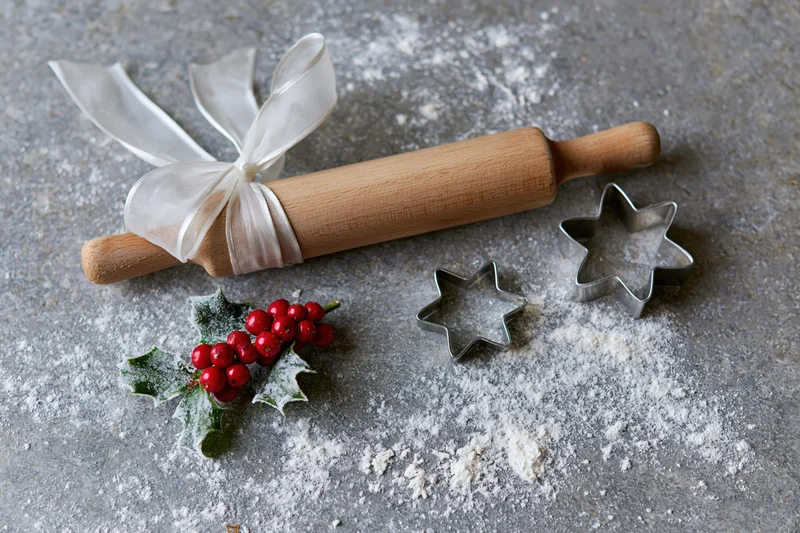 Wooden rolling pin tied with white ribbon and star-shaped cookie cutters on a floured gray countertop with holly berries, for holiday baking