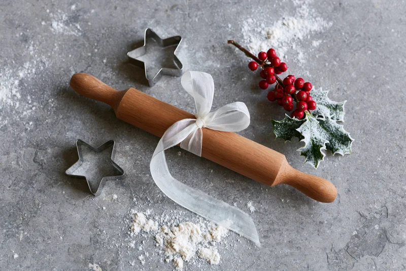 Wooden rolling pin tied with white ribbon beside star-shaped cookie cutters and holly sprig on floured gray surface