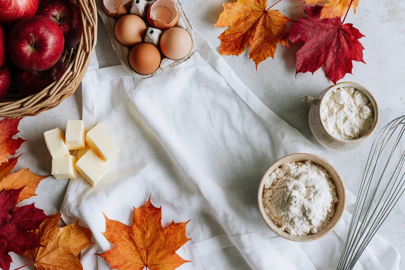 Baking ingredients arranged with autumn leaves on a white cloth on a kitchen countertop