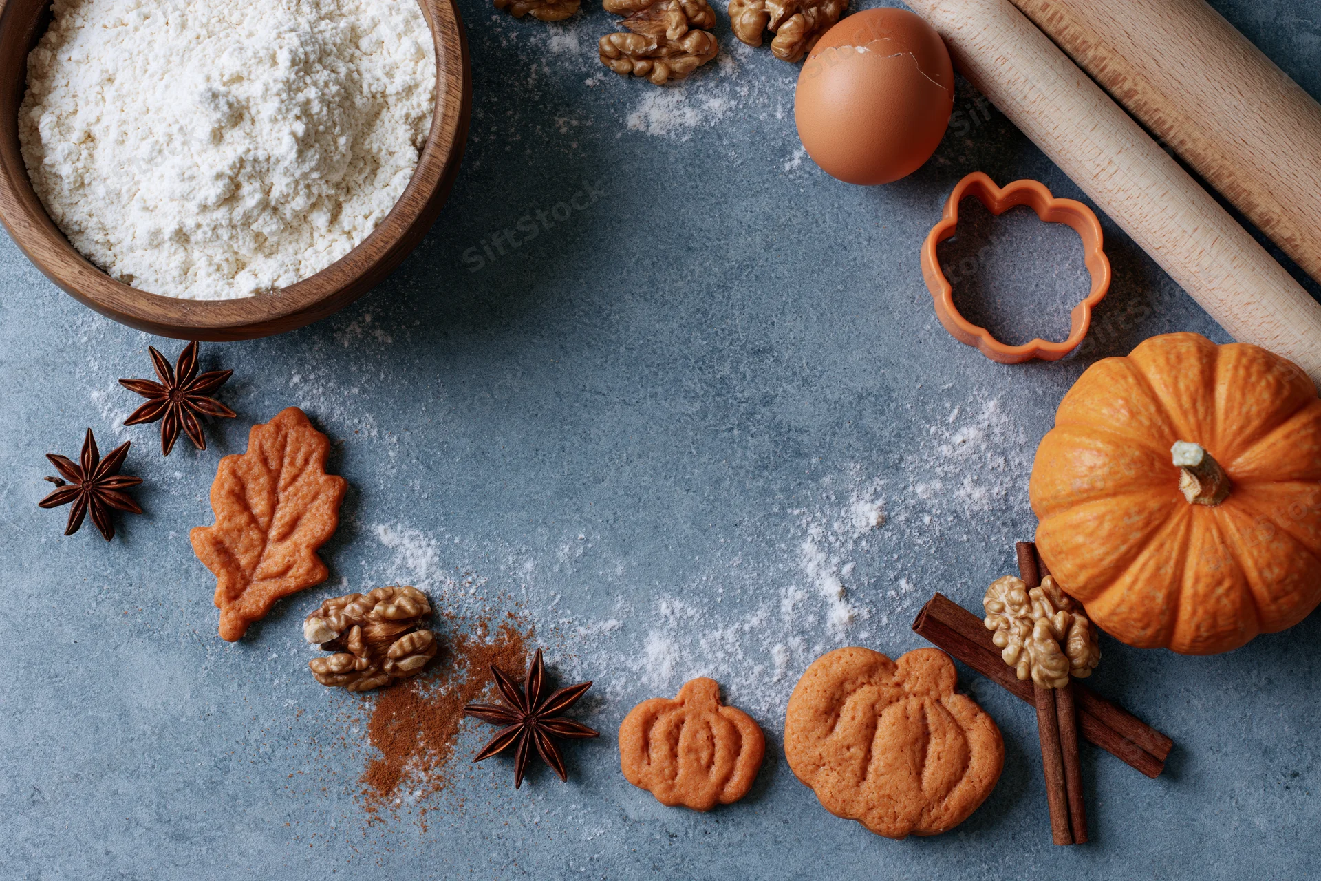 Pumpkin-shaped cookies and baking ingredients arranged on a gray countertop with autumn spices and a small pumpkin
