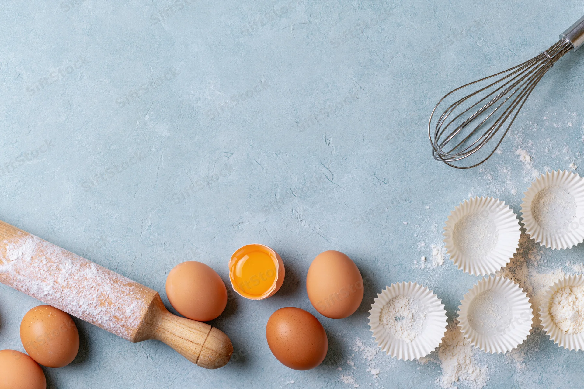 Baking ingredients arranged on a textured blue surface with a rolling pin, eggs, whisk and cupcake liners