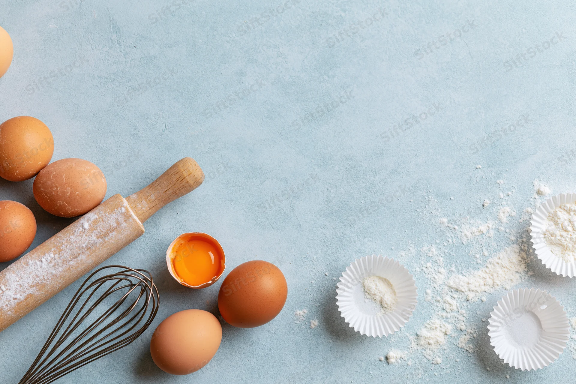 Brown eggs and baking tools arranged on a blue surface with flour and paper liners