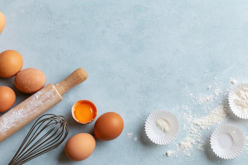 Brown eggs and baking tools arranged on a blue surface with flour and paper liners