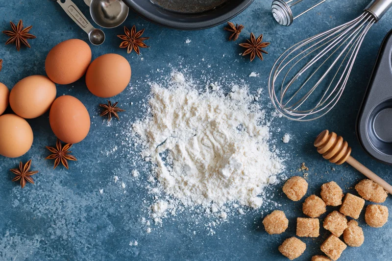 Baking ingredients arranged on a blue countertop with flour, eggs, sugar, star anise, whisk and honey dipper