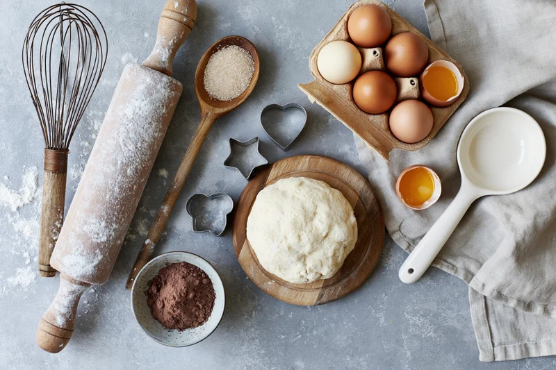Dough on a wooden board surrounded by baking tools and ingredients on a gray countertop